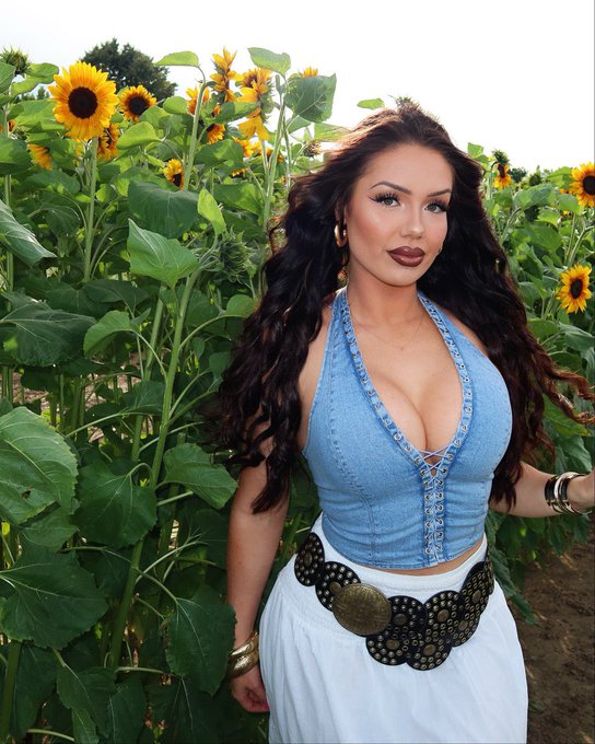 A woman with long dark hair posing in a sunflower field, wearing a denim halter top, white skirt, and a large decorative belt with coin details.