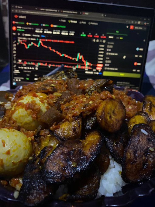 Plate of Indonesian food, likely nasi campur with tempeh oregan, eggs, meat, and sambal, with a laptop displaying a red/green stock or crypto chart in the background.
