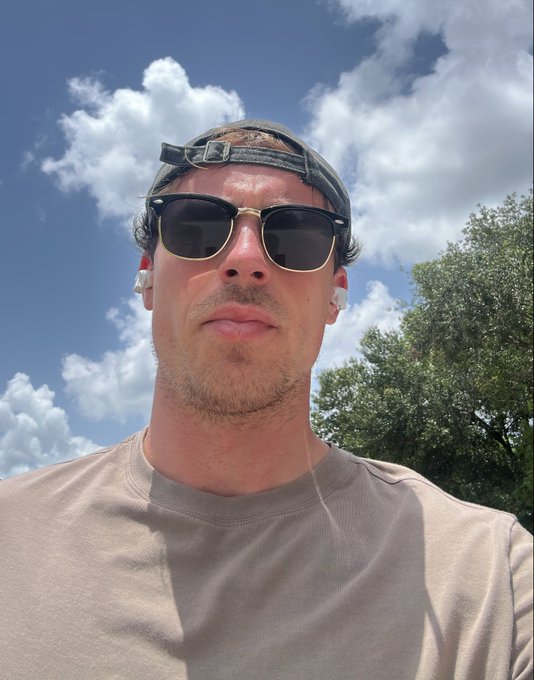 A selfie of a man wearing a backwards gray cap, dark sunglasses with a gold frame, white wireless earbuds, and a beige t-shirt, taken outdoors under a blue sky with clouds.