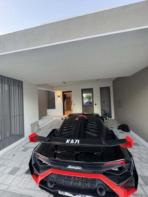 A glossy black and red Lamborghini, featuring "KA71" branding and a prominent rear spoiler, is parked under the covered entryway of a modern home, perfectly setting the tone for an exciting "Good Morning" ahead.