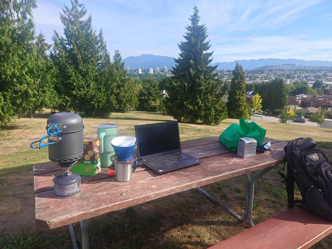 A portable coding setup with a laptop, backpacking stove, and coffee gear sprawled across a picnic table overlooks a scenic city and mountain view, suggesting the freedom of remote work in nature.