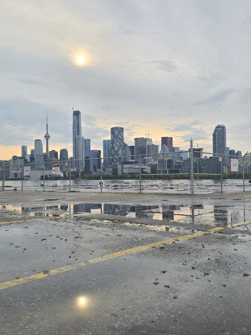 Toronto skyline at dusk viewed through a chain-link fence, with puddles reflecting the CN Tower and city buildings on a wet pavement.