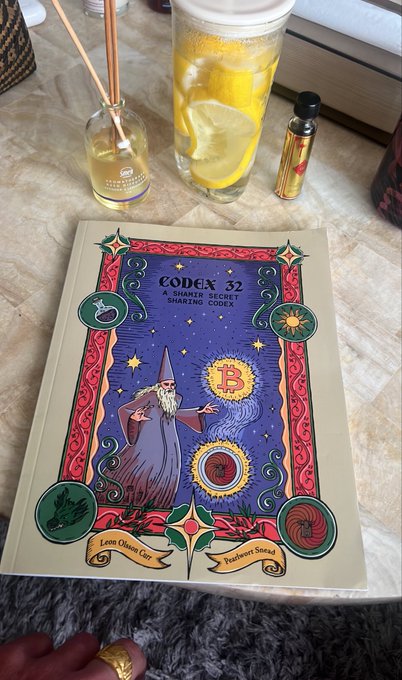 A hardcover book titled "CODEX 32: A SHARIAH-SECRET SHARING CODEX" with ornate artwork lies on a table next to a reed diffuser, a glass of lemon water, and a small spray bottle.