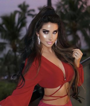 A woman with long dark hair wearing a deep V-neck red dress and statement earrings poses outdoors with palm trees visible in the background.