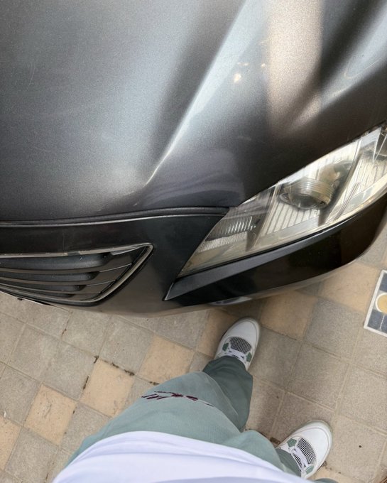 A person stands on a tiled sidewalk next to the front end of a dark gray car with a visible dent on the hood and a clear headlight, appearing to inspect or photograph the damage as part of a "gm" (good morning) greeting.