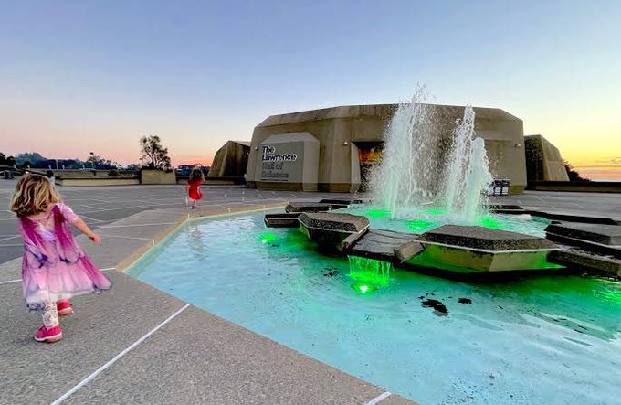 A young girl in a pink and purple dress standing near a fountain with green-lit water in front of a building labeled 'The Lawrence', at sunset.