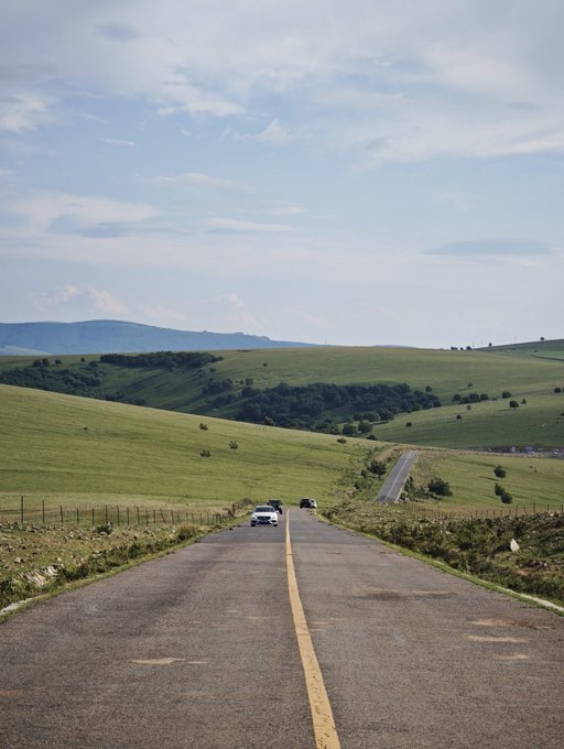 A scenic view of an empty country road stretching through green rolling hills under a partly cloudy blue sky, with a couple of cars driving in the distance.