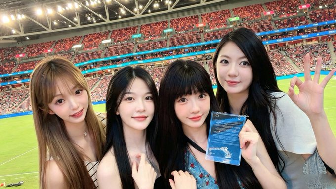 Four young women pose together for a selfie at a large sports stadium, with one holding a blue ticket or card while making a peace sign.