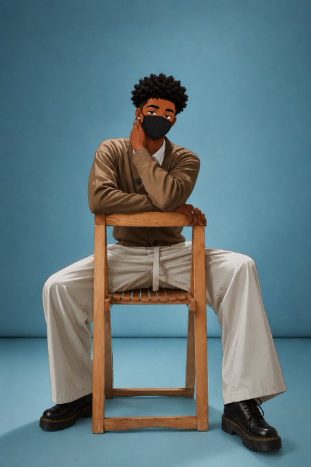 A studio portrait of a young man with curly hair wearing a brown cardigan, beige pants, black boots, and a black face mask, sitting on a wooden chair against a blue background.