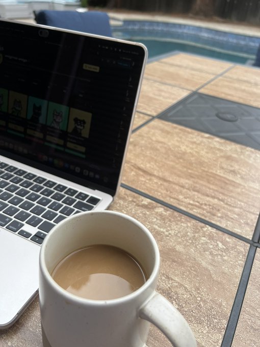 A MacBook laptop sits on a tiled outdoor table next to a white ceramic mug filled with coffee, with a swimming pool visible in the background.