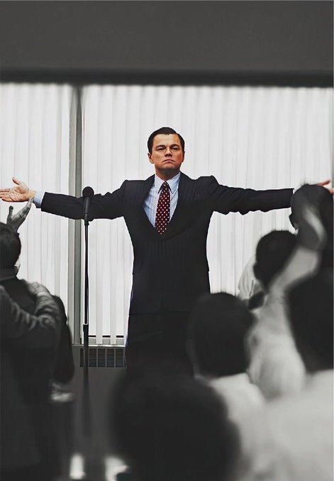 Leonardo DiCaprio stands with arms outstretched in a dramatic scene, dressed in a dark suit and red tie, addressing a seated audience with vertical blinds visible behind him.