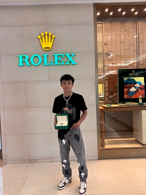 A young man stands in front of a Rolex storefront holding a green Rolex box, wearing a black graphic t-shirt and grey patterned jeans.