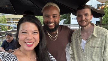 A selfie of three people smiling outdoors at what appears to be an outdoor venue with string lights visible in the background.
