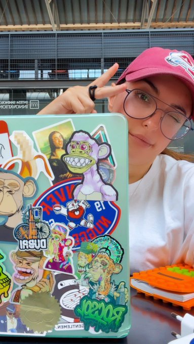 A woman wearing a pink cap and glasses poses beside a laptop heavily decorated with stickers, including a Mona Lisa and various logos.