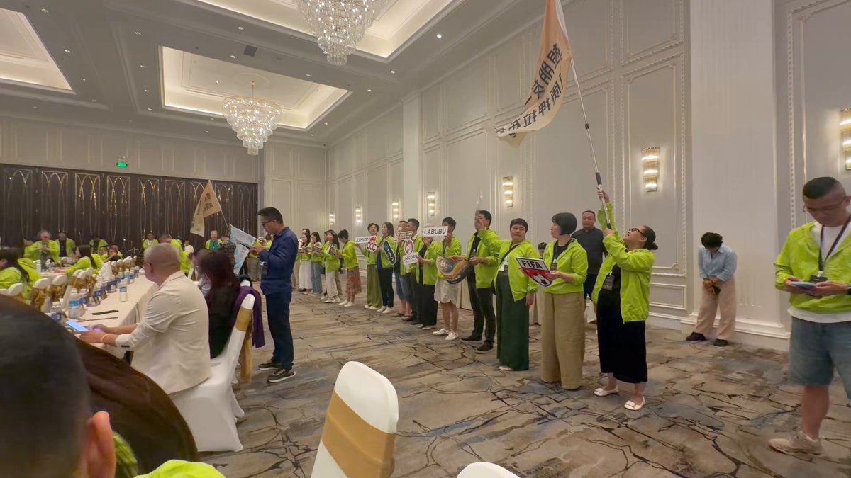 The image shows a group of people standing together, possibly at an event or gathering. They are wearing green shirts and holding up a banner with Chinese characters on it. The scene takes place inside a large room that appears to be a banquet hall or conference center. There is also a dining table visible in the background. A person can be seen sitting at one of the tables, possibly waiting for their turn to speak or participate in an activity.