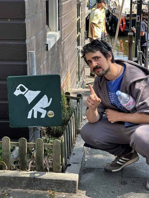 A man crouching next to a sign featuring a white dog illustration, pointing at it while holding a Vita water bottle, appearing to be near a canal or waterway.