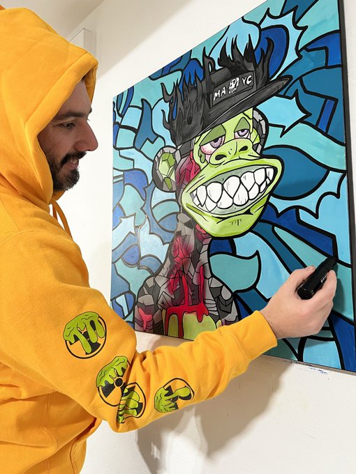 A man in a yellow hoodie with green smiley face designs photographs a colorful graffiti-style painting of a green character wearing a black cap labeled 'MADYO'.