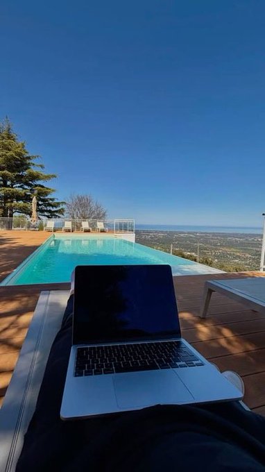 A person sits by an infinity pool overlooking a landscape, working on a laptop placed on their lap under a clear blue sky.