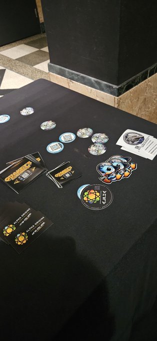 A black table displays a collection of crypto-related merchandise including holographic stickers, business cards, and patches featuring turtle and other designs.