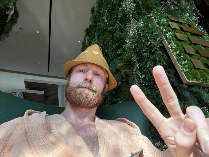 A bearded man wearing a yellow bucket hat and peach shirt takes a selfie while making a peace sign in front of a green plant wall.