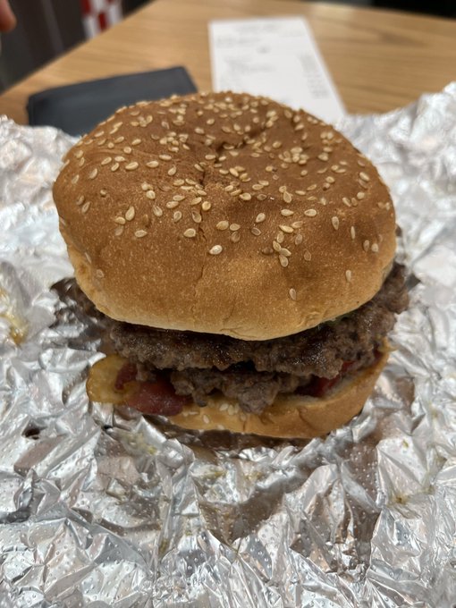 A close-up photograph of a sesame seed hamburger with a beef patty and visible sauce, resting on crinkled aluminum foil on a wooden table.