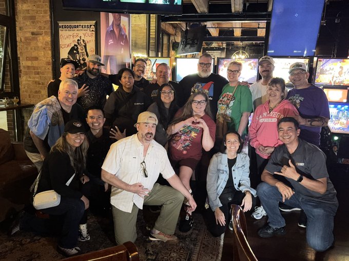 A large group photo of diverse people posing together at what appears to be an arcade or entertainment venue called 'HEADQUARTERS', featuring arcade machines in the background and a 'San Marcos Texas' t-shirt on one person.