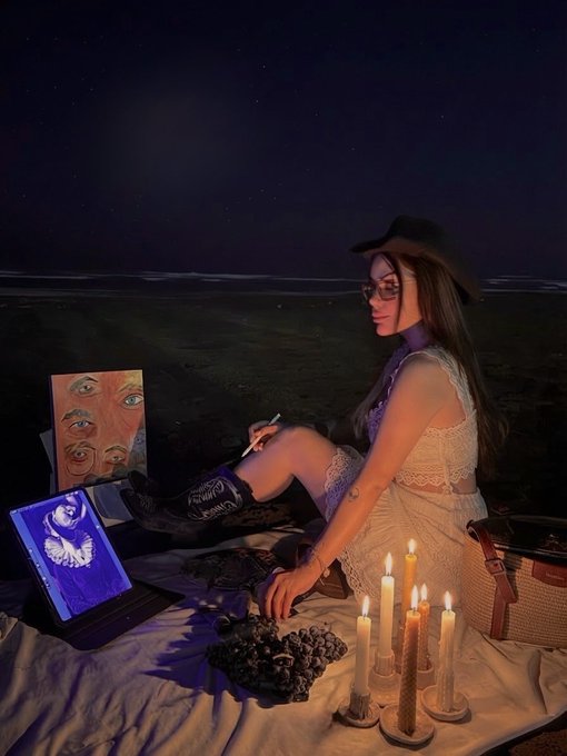 An atmospheric nighttime photo of a woman in a white dress and cowboy hat sitting by the ocean with candles, grapes, and a small screen displaying a blue figure.