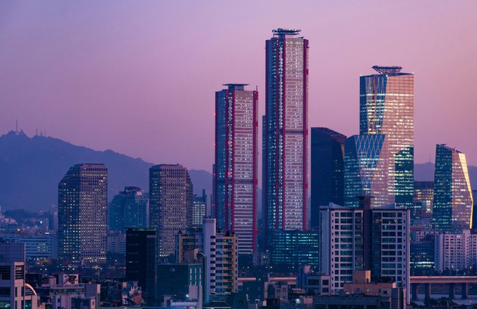A city skyline at dusk features multiple illuminated skyscrapers, including a tall tower under construction, set against a purple-pink sky with mountains visible in the background.
