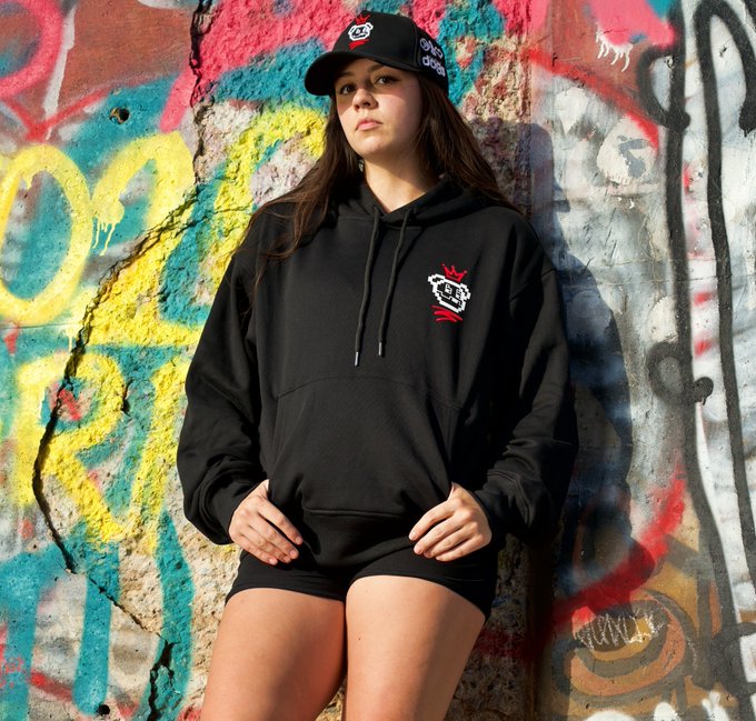 A young woman poses in front of a graffiti-covered wall, wearing a black hoodie and matching shorts that feature a small white logo.
