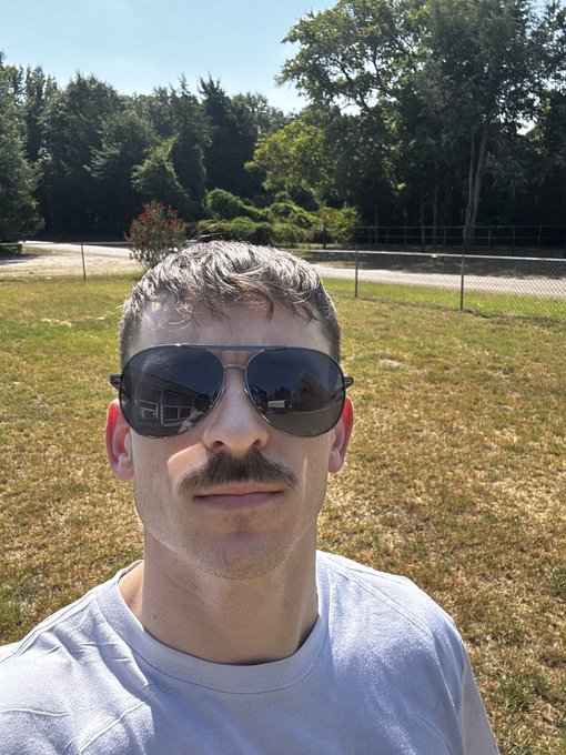 A man with a mustache and aviator sunglasses takes a selfie outdoors on a sunny day, with a grassy field, chain-link fence, and trees in the background. The image fits the post's motivational Monday theme, capturing a ready-to-go vibe as he faces the camera with a calm, determined expression.