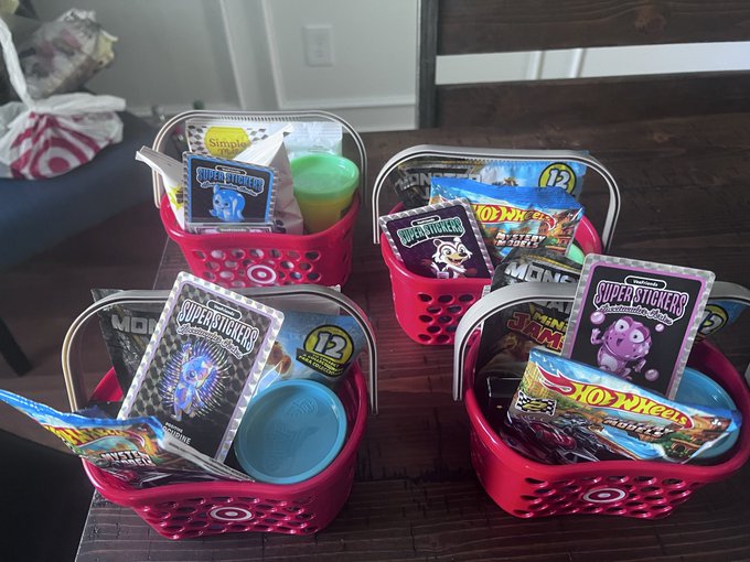 Four red Target shopping baskets filled with children's items, including 'SUPER STICKERS' trading cards, Hot Wheels cars, slime containers, and Play-Doh, sitting on a wooden table.