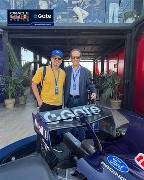 Two men wearing lanyards stand beside a Red Bull Racing F1 car marked "Oracle," "Gate," and "Ford Racing" at the Red Bull Burladero Hospitality, capturing the celebratory atmosphere of Max Verstappen's P2 podium finish and wishing Isack Hadjar a speedy recovery.