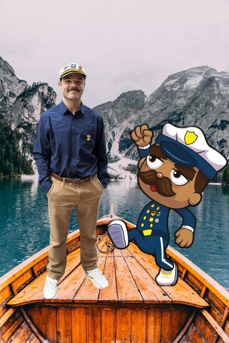 A man wearing a blue button-down shirt, khaki pants, and a captain's hat stands on a wooden boat on a mountain lake, posed next to a cartoon police officer character sticker.