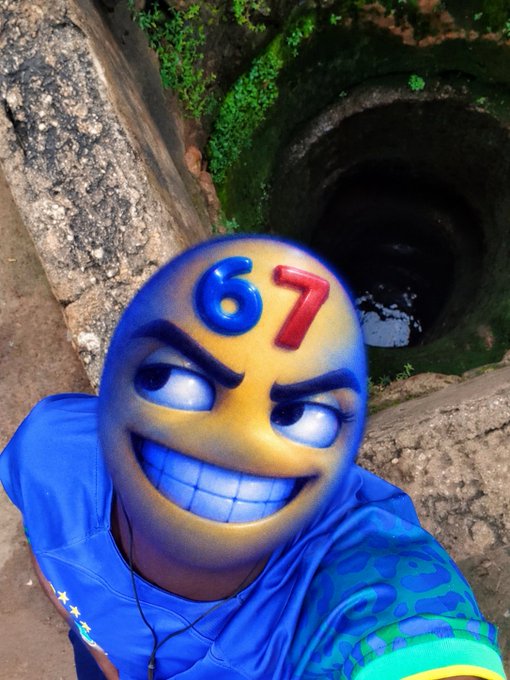 A selfie of a person in a blue shirt with a digitally applied '67' emoji-style face mask, standing near a stone well with green moss.