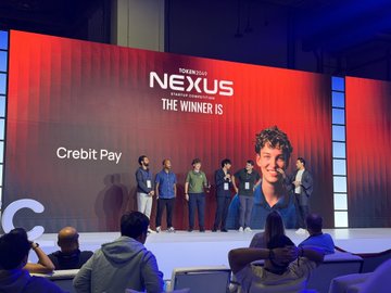 A stage event displaying 'NEXUS' and 'THE WINNER IS Crebit Pay' on a large red screen, with a group of people standing on stage in front of it.