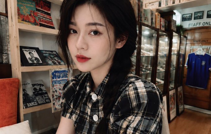 A young woman with dark hair wearing a plaid shirt posing indoors in front of wooden shelves displaying vintage magazines and records.