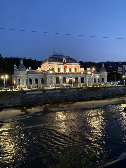 A grand, illuminated historic building stands majestically along a riverbank at dusk, its architectural details and warm lights reflecting beautifully on the water below, capturing the charm of Eastern European heritage. The serene nighttime scene complements the post's theme, pairing the timeless beauty of the region with a modern countdown toward ENA's $3 target on day 642.