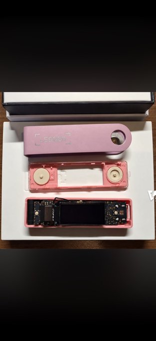 A pink Ledger hardware wallet is shown disassembled in its white box, displaying the device case, internal components with two button cells, and the circuit board.