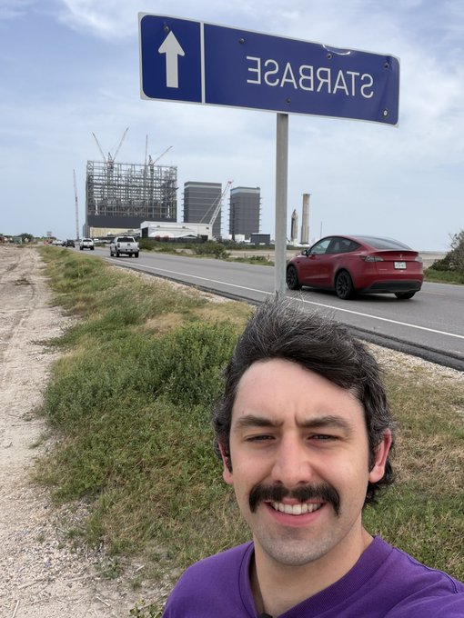 A selfie shows a smiling man with a mustache in a purple shirt, taken below an upside-down 'STARBASE' road sign with industrial buildings visible in the background