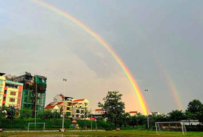 A double rainbow arcs over a residential area with apartment buildings, a soccer field, and a sign reading 'BUV', under a partly cloudy sky.