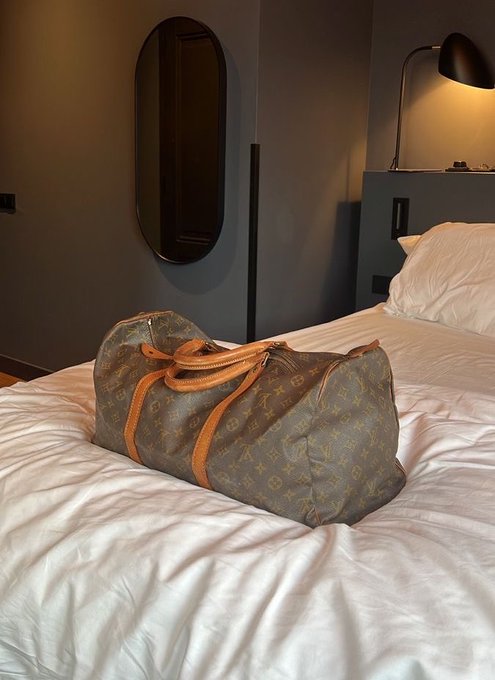 A brown Louis Vuitton monogram Keepall duffle bag rests on a white-sheeted bed in a dimly lit hotel room with a modern lamp.
