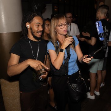 Two people—a man with earphones and a woman with glasses—sing karaoke in a dimly lit venue while someone in the background records them on a smartphone.