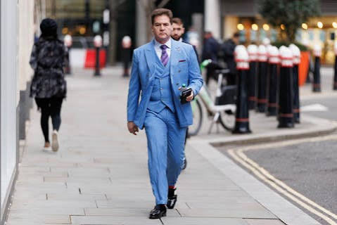 A man in a bright blue three-piece suit walks down a city sidewalk, holding a phone, with red bollards and a bicycle visible behind him.
