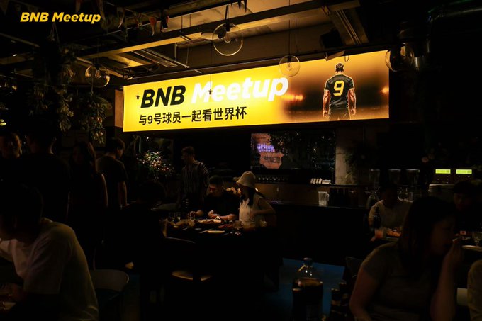 A dimly lit venue hosts a "BNB Meetup" event featuring a large illuminated sign reading "BNB Meetup 与9号球员一起看世界杯" alongside an image of a player wearing jersey number 9, where attendees gather around tables likely to celebrate Binance's anniversary and watch the World Cup.