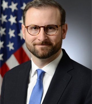 An official portrait of a bearded man wearing glasses, a dark suit, and blue tie, standing in front of an American flag.