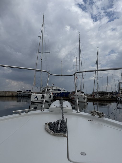 A view from the bow of a white sailboat moored in a harbor, looking out at several other sailboats and yachts docked under a cloudy sky.