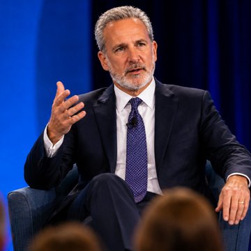 In this image, a man dressed professionally, wearing a suit and tie, is seated comfortably on a blue chair inside a large room with a blue background, suggesting an indoor setting such as a conference or presentation hall. The central focus of the image is the man's hands placed near his mouth, which he appears to be gesturing with during the speech. 

The image also mentions Peter Schiff and Bitcoin, indicating that this could potentially be related to financial discussions. Schiff warns that if Bitcoin falls below $50,000, it might reach a new low in value for investors who have been holding Bitcoin, possibly impacting their overall portfolio returns or gains they've already achieved from Bitcoin ownership.