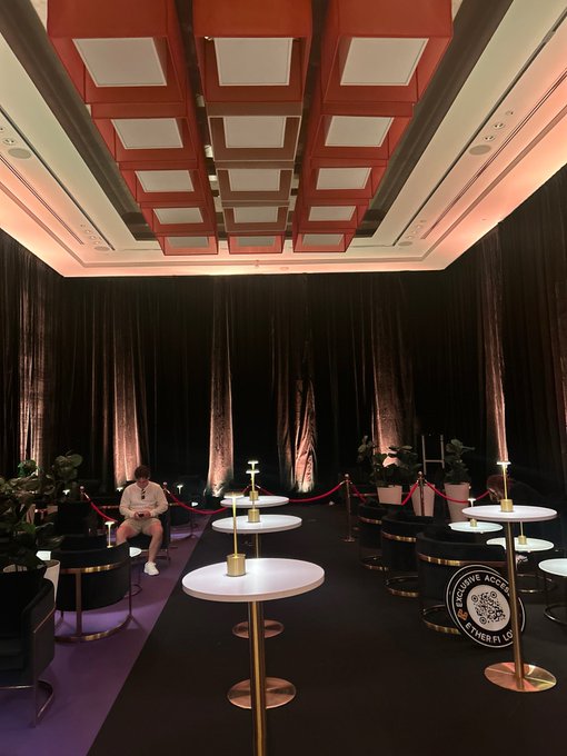 An elegant lounge setup featuring small round cocktail tables, dark velvet seating, and decorative red ceiling panels awaits attendees of tomorrow's @ether_fi event, where attendees are invited to pick up their cards. The watermark "Genius Access & ether.fi" is visible on one of the tables, tying the sophisticated venue to the upcoming gathering.