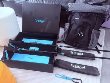A black 'Bitget' branded box containing a small cyan tripod, with additional Bitget-branded tripod bags and accessories laid out on a bed.