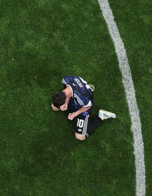 An aerial view of a soccer player in a dark jersey with the number 10 kneeling on a green field near the white sideline, appearing to kiss his hand.